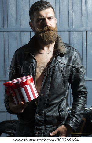 Handsome unshaven young man with beard and moustache in brown leather jacket and chain holding round red white striped present box with ribbon bow standing on grey wooden background, vertical picture