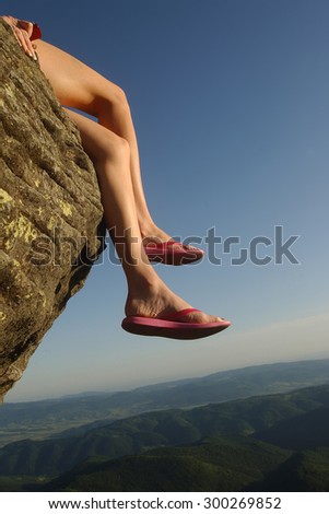 Beautiful view from high mountaing on green hills with forest and human female legs in slippers of woman sitting on rock on natural blue sky space background, vertical picture