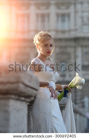 Pretty sensory young blond fiancee with stylish hairdo in white wedding dress holding bouquet of calla flowers with blue ribbon standing on stone bridge outdoor sunny day, vertical picture