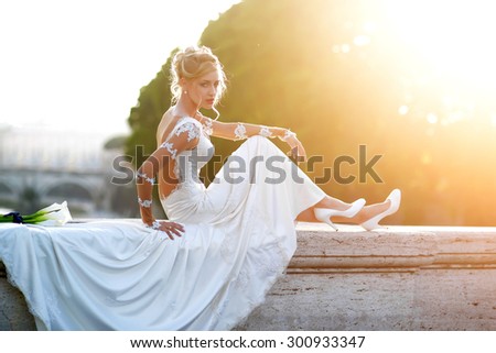 Pretty young fiancee with elegant hairdo in white wedding dress with long train and shoes sitting on stone bridge parapet with bunch of calla flowers with blue ribbon outdoor, horizontal picture