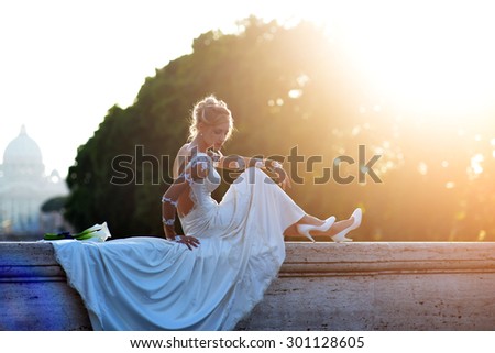 Attractive sensual bride with elegant hairdo in white wedding dress with long train and shoes sitting on stone bridge parapet with nosegay of calla flowers with blue ribbon outdoor, horizontal picture