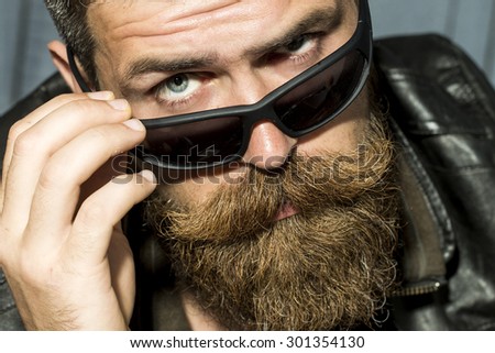 Portrait of serious brutal unshaved male biker with long beard and moustache in brown leather jacket and black sunglasses looking forward closeup, horizontal picture