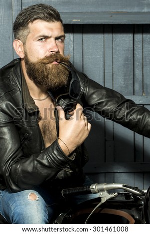 Attractive brutal unshaven sexy male biker in brown leather jacket jeans with sunglasses sitting in garage on motor cycle looking forward on wooden background, vertical picture