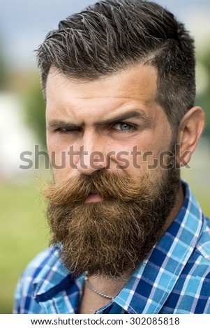 Portrait of handsome young unshaven man with long beard and hendlebar moustache in checkered white and light blue shirt looking forward standing outdoor, vertical picture