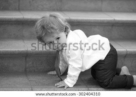 Little curious stylish baby boy with curly hair in formal shirt necktie and trausers creeping barefoot on stone stairs looking away black and white, horizontal picture