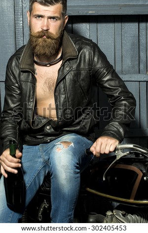 Handsome sexy brutal male biker with long beard and moustache in brown leather jacket with open chest and blue jeans holding glass wine bottle looking forward sitting on motor cycle, vertical picture