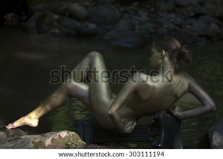 Beautiful stripped woman mermaid with sensual painted gold body lying with back on stony rock coast in wood near water sunny day outdoor on natural background, horizontal picture