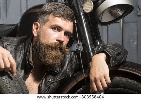 Young unshaven male biker in leather jacket sitting near motorcycle in garage with big black rubber spare wheel looking forward on wooden wall background, horizontal picture