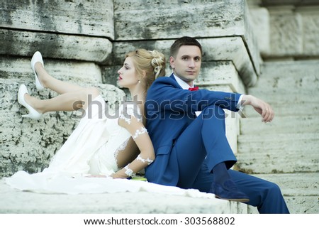Beautiful young wedding couple of sensual blond girl in white dress and shoes and man in blue suit with red bow tie sitting near stone building outdoor, horizontal picture