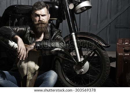 Sullen unshaven male biker in leather jacket sitting near motorcycle in garage with big bone skull antlers of stuffed animal looking forward on wooden wall background, horizontal picture