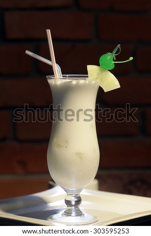 Glass with alcoholic pina colada cocktail of light rum coir coconut milk crushed ice frappe pineapple juice and slice green cherry and drink straws on white plate on brick background, vertical photo