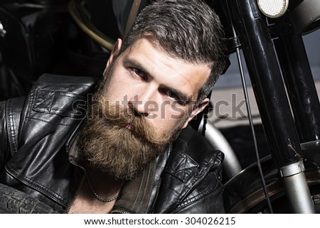 Portrait of handsome pensive unshaven man with beard and handlebar moustache in black leather biker jacket sitting near motorcycle looking forward, horizontal picture