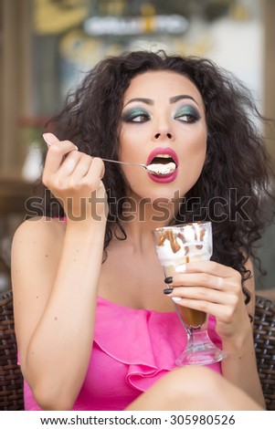 Portrait of sexual pretty brunette lady with curly hair and bright makeup eating cold dessert of ice cream and coffee glissade from glass with spoon, vertical picture