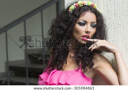 Beautiful sexy brunette young woman with wreath of colorful flowers in curly hair and bright makeup in pink clothes standing near white wall and stairs looking away, horizontal picture