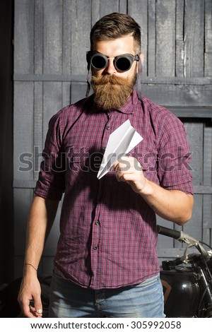 Attractive unshaven male biker in purple checkered shirt and glasses standing near motorbike in garage holding white paper plane looking forward on workshop background, vertical picture