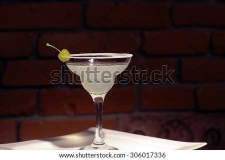 Glass with alcoholic white lady cocktail of lemon juice liqueur cointreau gin and green cherry with sugar on rim of glass on white plate on brick wall background, horizontal photo