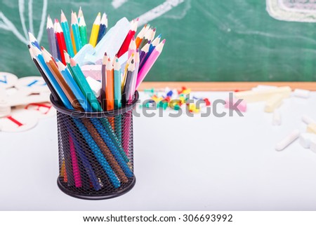 Colorful pencils of red yellow orange violet purple pink green and blue in stationary cup on school desk with white sheet of paper on written with chalk blackboard background, horizontal picture