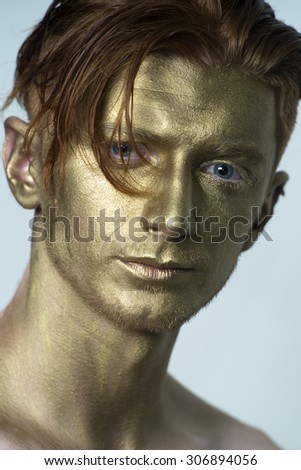 Portrait of young fashionable painted man model with golden bodyart on face and stylish red hairdo looking forward standing in studio on white background, vertical picture