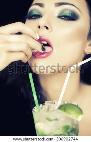 Portrait of cool pretty brunette woman with curly hair and bright makeup with alcoholic mojito cocktail from mint soda light rum and lime with straw licking ice cube, vertical picture