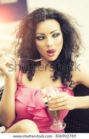 Sexual playful brunette lady with curly hair and bright makeup in pink clothes eating cold dessert of ice cream and coffe glissade from glass with spoon, vertical picture