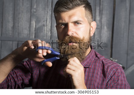 Portrait of serious man in violet checkered shirt cutting long beard and handlebar moustache with scissors making new style looking forward standing on wooden wall background, horizontal picture