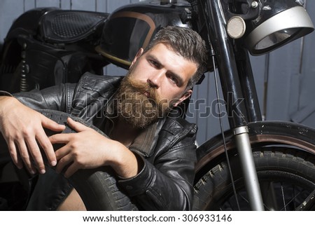 Attractive unshaven male biker in leather jacket sitting near motorcycle in garage with big black rubber spare wheel looking forward on wooden wall background, horizontal picture