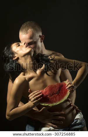 Young sensual undressed couple of pretty tan girl in jeans shorts holding big juicy red water melon slice and man with strong muscular beautiful body standing on black background, vertical picture