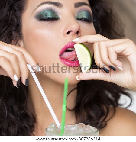 Portrait of young pretty brunette woman with curly hair and bright makeup with alcoholic mojito cocktail from mint soda light rum ice cubes with straw licking lime with closed eyes, square picture