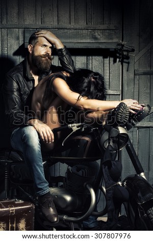 Young sexy couple of unshaven pensive biker in leather jacket and jeans with undressed brunette woman sitting together on motorcycle in garage on wooden wall background, vertical picture