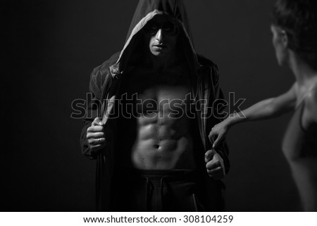 Young couple of man with sexy strong muscular beautiful body in leather jacket with hood and sunglasses and undressed woman standing in studio black and white, horizontal picture