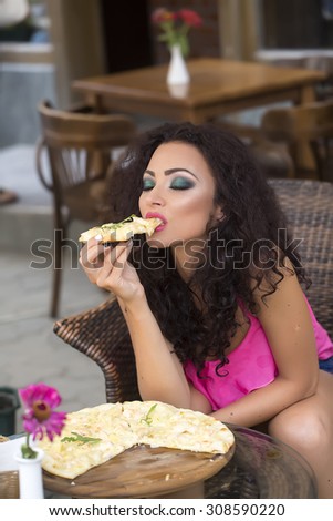Sensual young brunette lady with curly hair and bright makeup in pink clothes sitting in cafe outdoor eating hot delicious pizza with hands, vertical picture