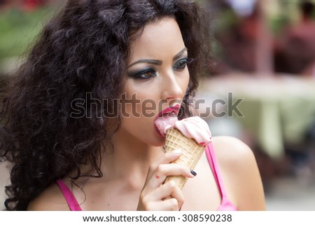 Portrait of sexy young brunette lady with curly hair and bright makeup licking cold dessert of red berry ice cream cone with tongue and open mouth, horizontal picture