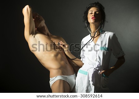 Sensual young pair of bare man in pants with strong muscular beautiful body and pretty brunette nurse in white smock with stethoscope standing on black studio background, horizontal picture