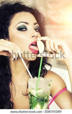 Portrait of young pretty brunette woman with curly hair and bright makeup with alcoholic mojito cocktail from mint soda light rum ice cubes with straw licking lime, vertical picture