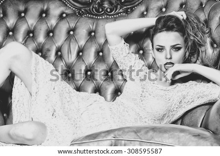 Attractive confident young stylish woman in lace dress with bright makeup looking forward lying on leather royal sofa indoor black and white, horizontal picture