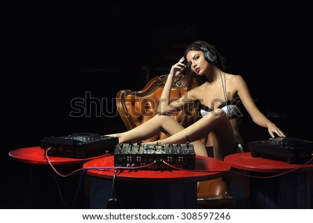 Beautiful young glamour dj woman in lingerie and headphones sitting at red table with mixer console on brown leather royal chair in night club on dark background, horizontal picture