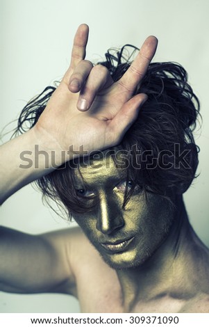 Portrait of young fashionable painted man model with golden bodyart on face and stylish hairdo holding hands in cool gesture looking forward standing in studio on white background, vertical picture