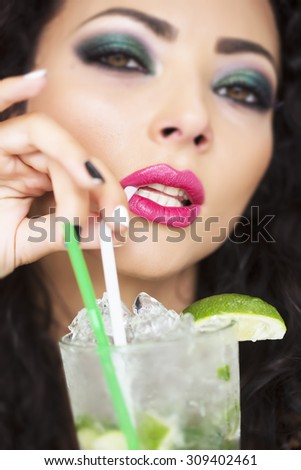 Closeup of sexy young brunette lady with curly hair and bright makeup drinking alcoholic mojito cocktail from mint soda light rum ice cubes and lime with straw, vertical picture