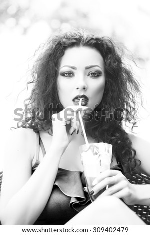 Portrait of sexual cute brunette lady with curly hair and bright makeup drinking cold dessert of ice cream and coffee glissade from glass with straw black and white, vertical picture