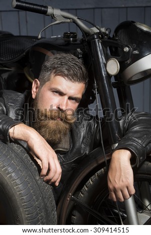 Young serious male biker in leather jacket sitting near motorcycle in garage with big black rubber spare wheel looking forward on wooden wall background, vertical picture