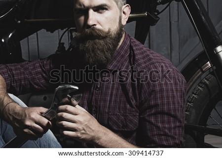 Young unshaven male biker in purple checkered shirt sitting near motorcycle in garage holding metallized iron wrench looking forward on workshop background, horizontal picture