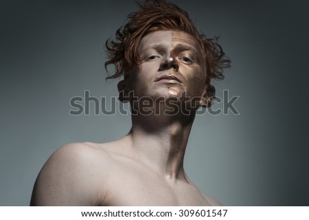 One young fashionable painted man model with bronzy bodyart on one half of face and stylish red hairdo looking forward standing in studio on grey background, horizontal picture