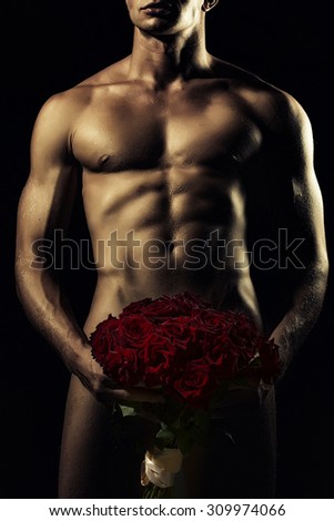 Young sexy undressed man with beautiful strong muscular body holding big fresh red rose flowers bouquet on genitals standing on black background, vertical picture