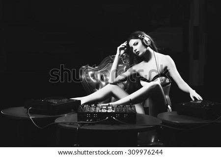 Pretty young glamour dj girl in underwear and headphones sitting at table with mixer console on brown leather royal chair in night club on dark background black and white, horizontal picture