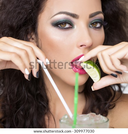 Portrait of young pretty brunette woman with curly hair and bright makeup with alcoholic mojito cocktail from mint soda light rum ice cubes with straw licking lime looking forward, square picture