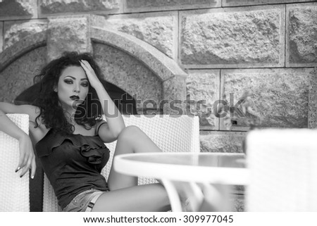 Sexy busty pretty brunette girl with curly hair and bright makeup in summer clothes sitting in cafe in chair at table with vase of fresh marigold flowers black and white copyspace, horizontal picture