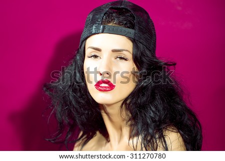 Portrait of young cool sexy sensual beautiful bad girl with brunette curly hair red glossy lips and black cap looking forward on bright pink studio background, horizontal picture