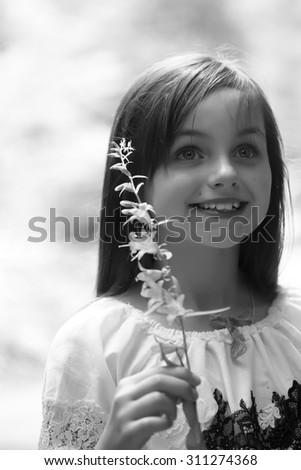 Portrait of pretty small smiling girl with long hair in blouse holding one branch of field bellflower bloom looking away standing sunny day outdoor black and white, vertical picture