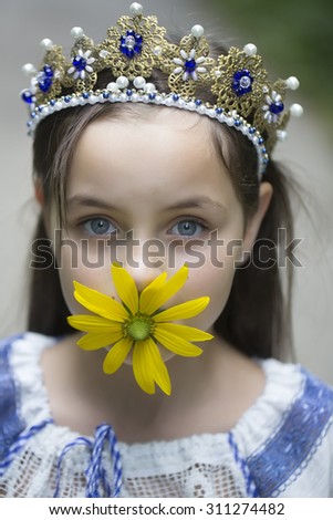 Portrait of pretty brunette princess girl in traditional ukrainian embroidery white blue colors and beautiful diadem with diamonds in gold holding yellow flower in mouth outdoor, vertical picture