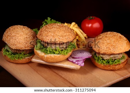 Big tasty appetizing fresh burgers of green lettuce red tomato cheese and bacon slice meat cutlet and white bread bun with sesame seeds chips and violet onion on black background, horizontal picture